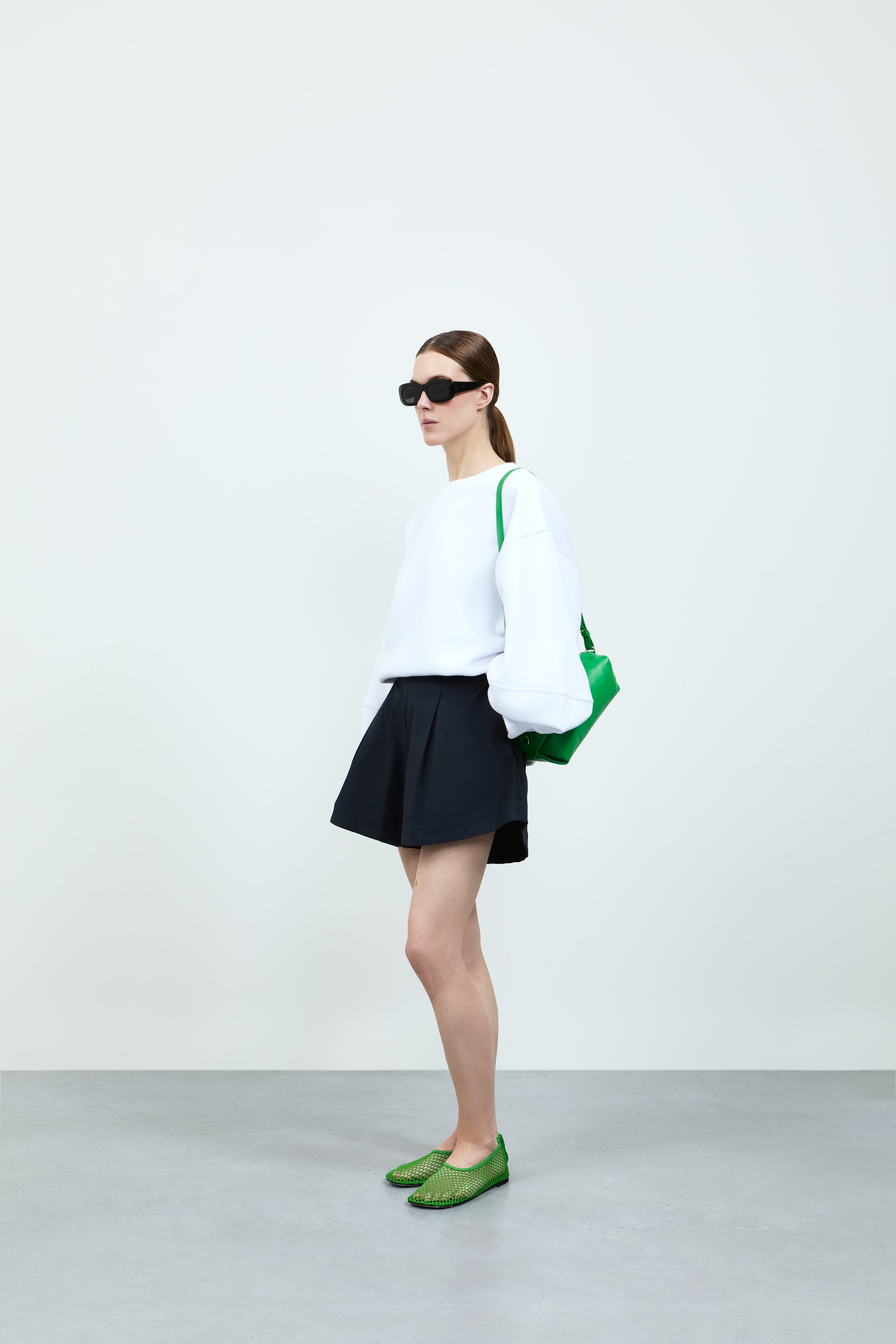 Person wearing a white sweater, black short, and green shoes on a plain background